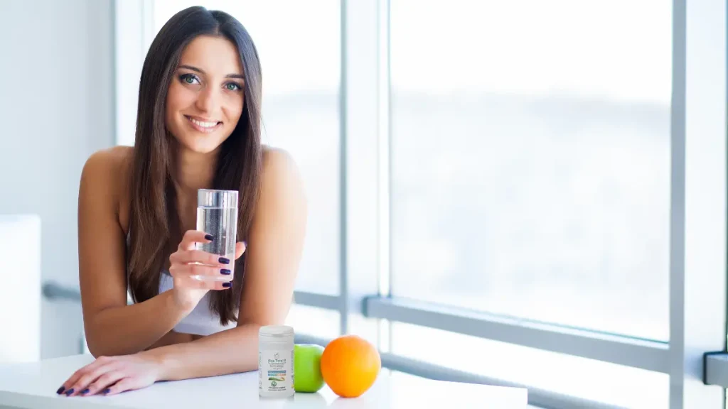 ragazza sorridente con bicchiere di acqua in mano con braccio appoggiato sul tavolo su cui ci sono frutta e vitamine del gruppo b